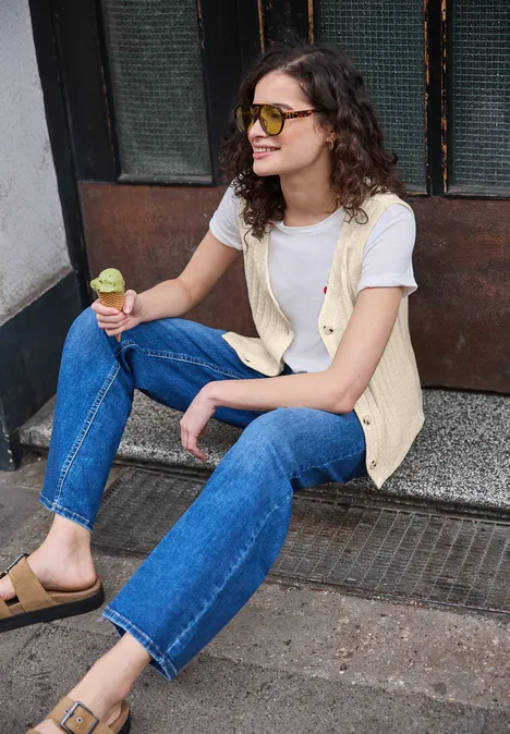 Person in Jeans, weißem Shirt und gelber Weste sitzt auf einer Schwelle und hält ein Eis in der Hand.